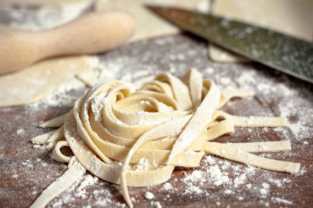 Freshly made pasta noodles dusted with flour are neatly arranged in a swirl on a wooden surface. A rolling pin and a knife sit nearby, suggesting a cooking or preparation scene.