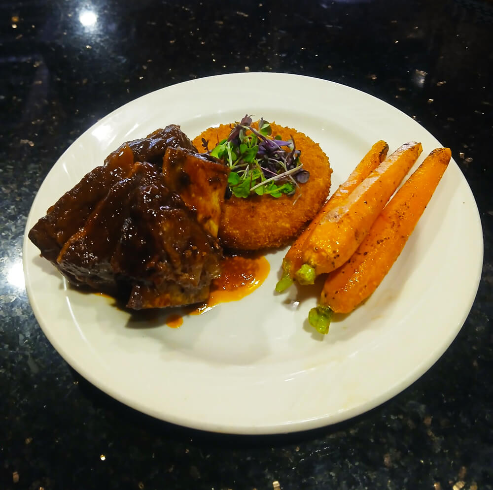 A white plate with braised ribs covered in sauce, garnished with microgreens, a crispy round potato cake, and three roasted carrots. It rests on a dark, glossy surface.