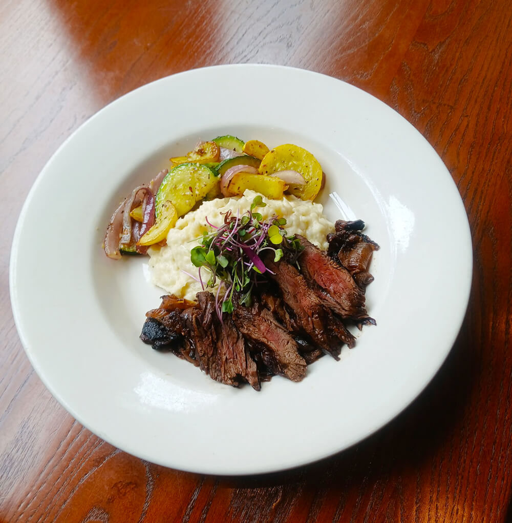A white plate with sliced grilled steak, creamy mashed potatoes topped with microgreens, and sautéed vegetables, including yellow squash, zucchini, and red onions, placed on a wooden table.