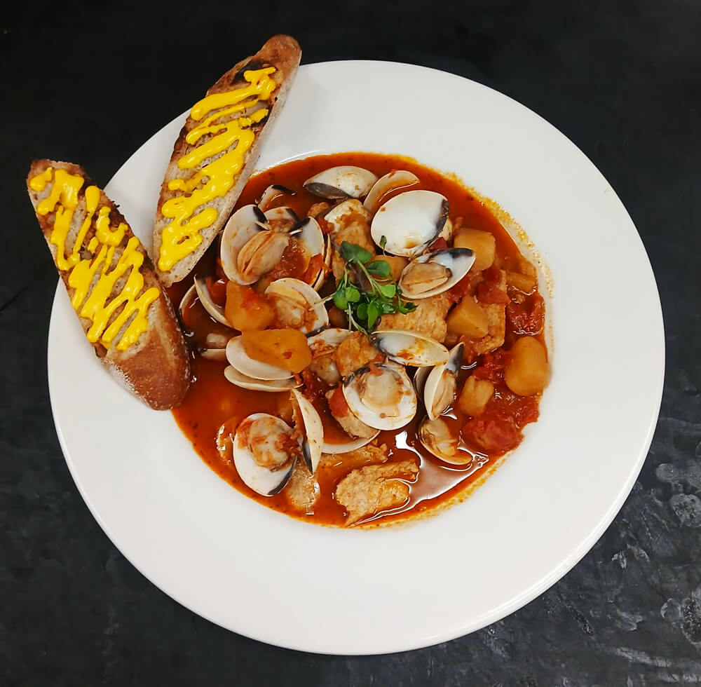 A white plate with a rich seafood stew containing clams, potatoes, and tomato broth, garnished with herbs. Two slices of crusty bread drizzled with yellow sauce are placed on the side. The background is a dark surface.