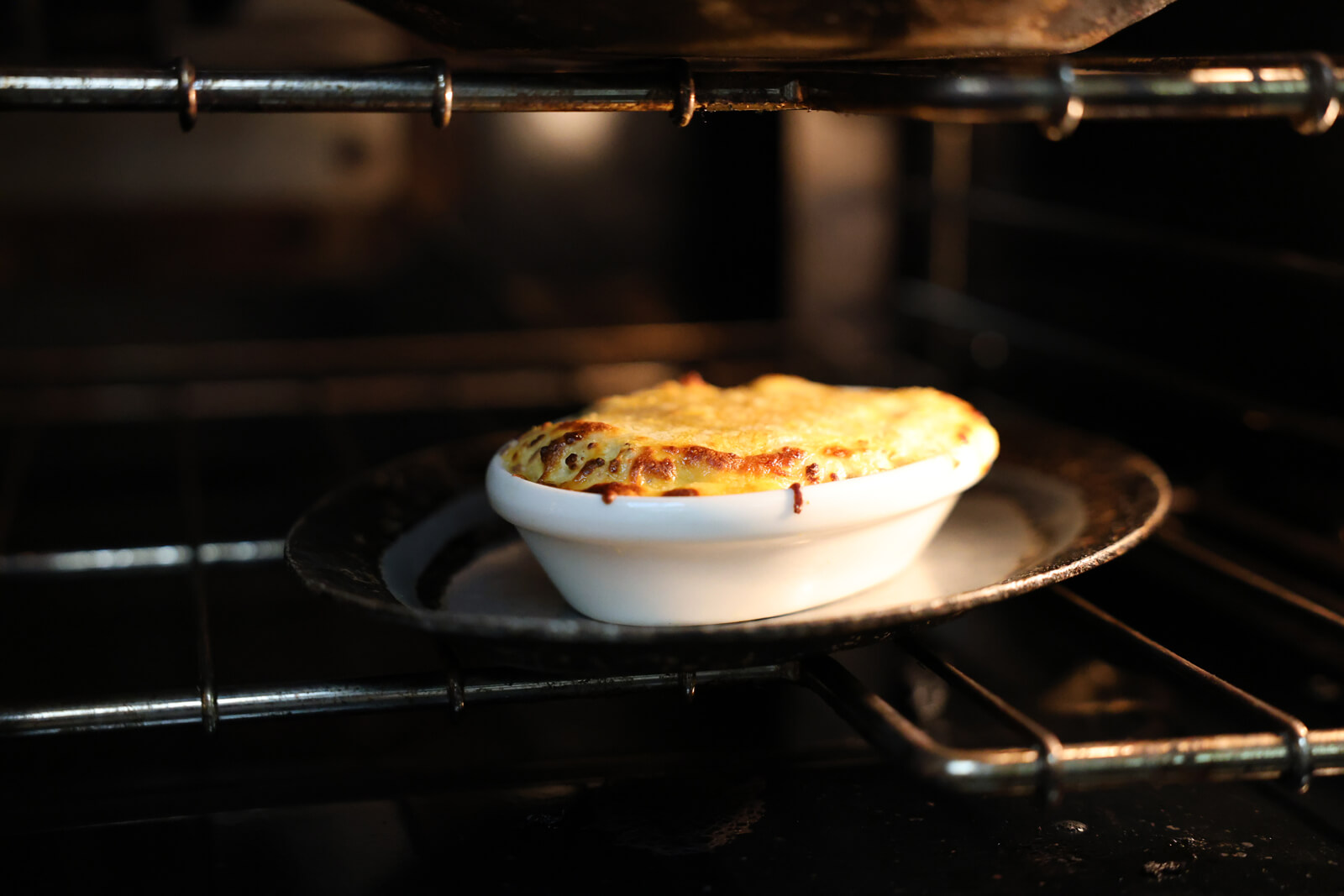 A small white dish filled with a baked, golden-brown casserole sits on a black tray inside an oven, with melted cheese bubbling on top.
