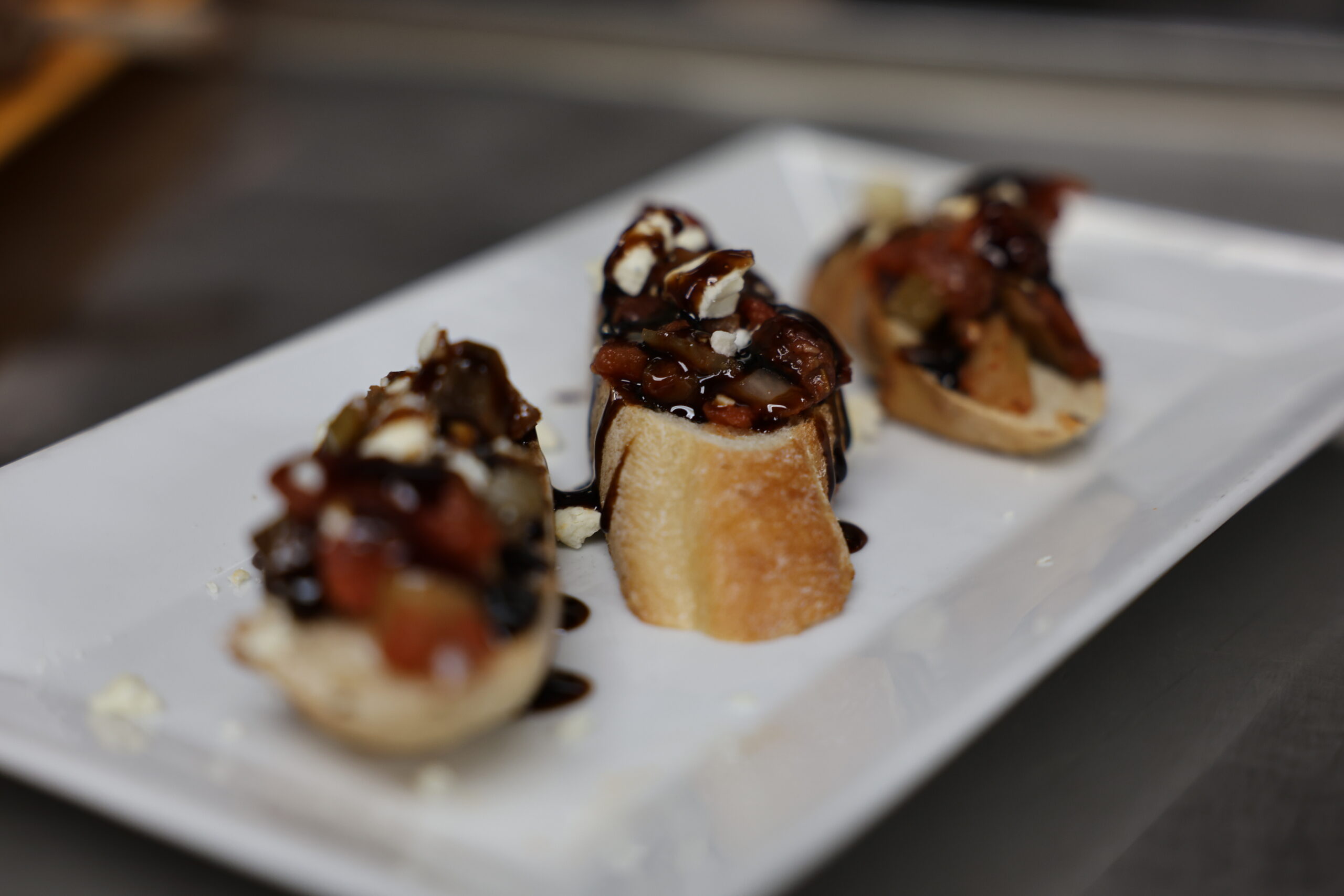 Four pieces of bruschetta topped with diced tomatoes, feta cheese, and balsamic glaze are arranged on a rectangular white plate.