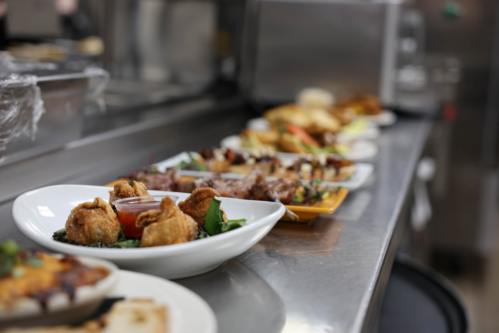 A row of plated food dishes sits on a stainless steel kitchen counter, with various appetizers, entrees, and garnishes lined up in a professional restaurant setting.