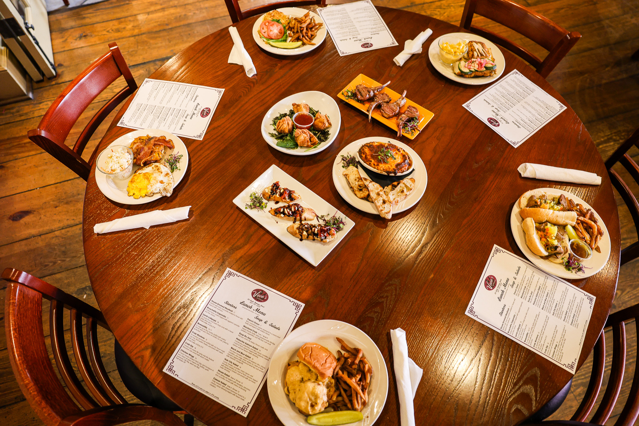 A round wooden table set with seven plates of assorted food, including sandwiches, fries, and salads, along with four printed menus and folded napkins on wooden chairs in a cozy restaurant setting.