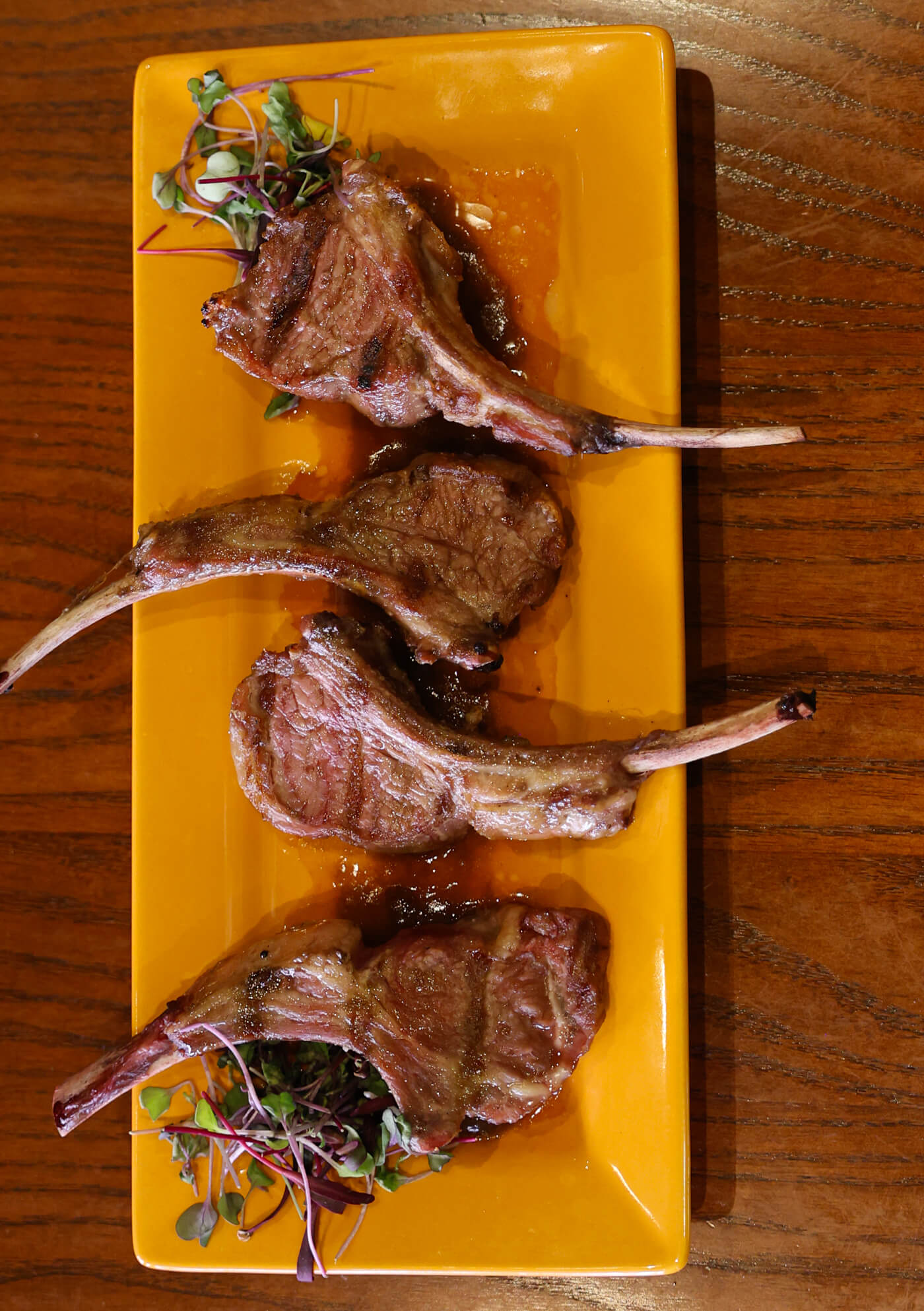 Four grilled lamb chops arranged on a rectangular yellow plate, garnished with microgreens, served on a wooden table.