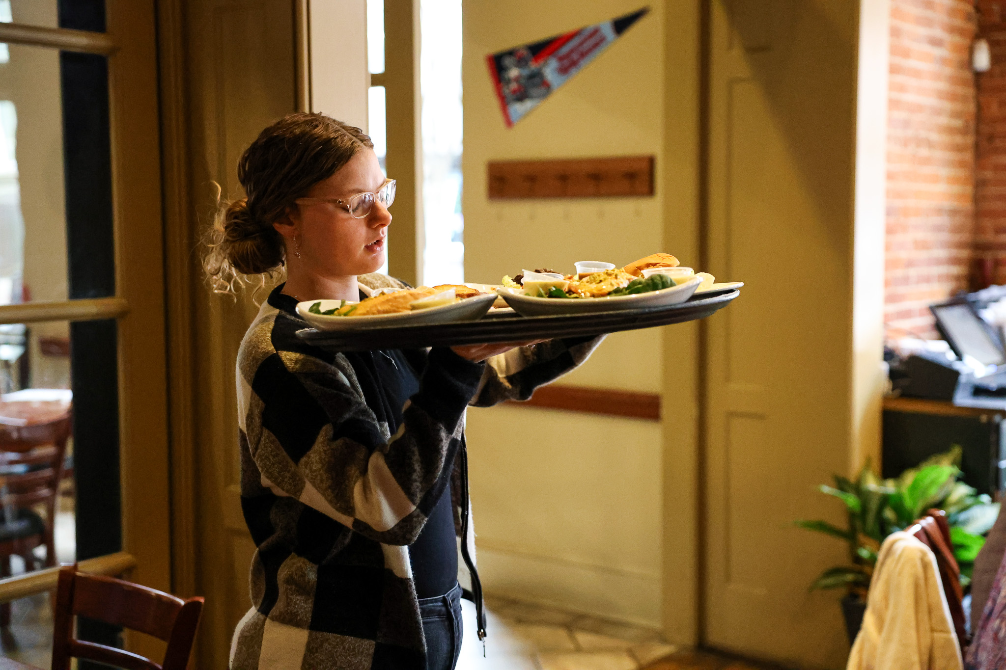 A server wearing glasses and a checkered sweater carries a tray with several plates of food through a restaurant. The background shows tables, chairs, a plant, and a wall with a pennant and coat hooks.