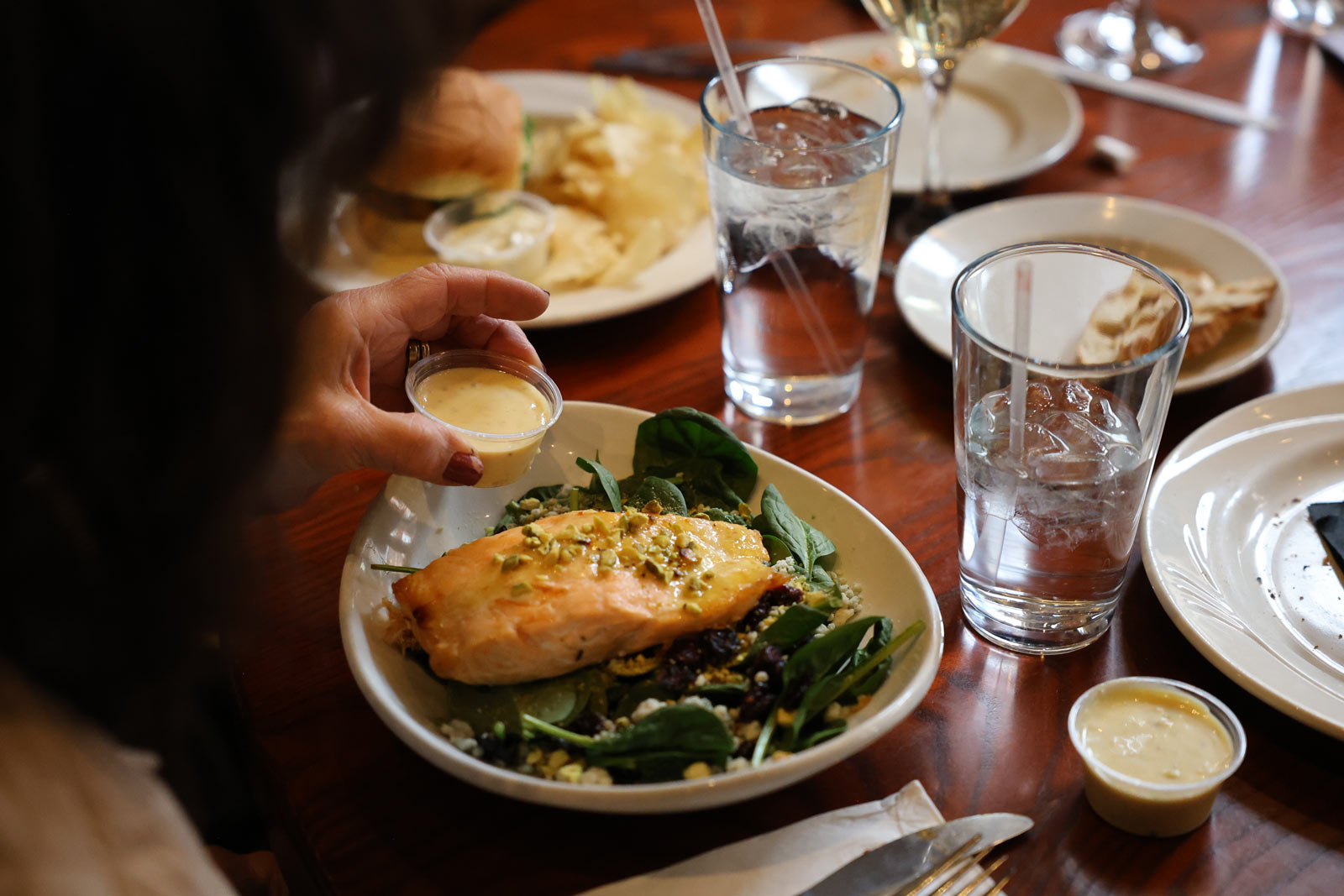 A person pours dressing onto a spinach salad topped with cooked salmon and nuts. The table holds plates, glasses of water, a glass of white wine, and various small dishes.