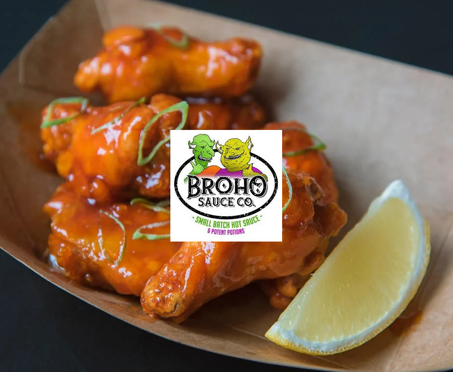 Buffalo chicken wings coated in a bright orange sauce, garnished with green herbs, served in a paper tray with a lemon wedge. A Broho Sauce Co. logo is centered over the wings.