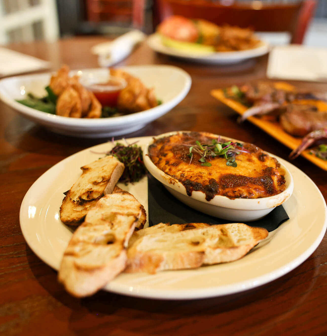 A plate of toasted bread slices next to a baked dish topped with melted cheese, garnished with microgreens. In the background, plates of fried appetizers and grilled meat are slightly out of focus.