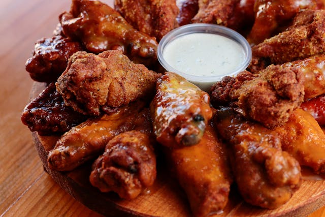 A wooden platter filled with assorted sauced and fried chicken wings arranged in a circle around a cup of creamy dipping sauce.